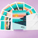 A set of illustrated postcards and white envelopes arranged on a lavender background. The postcards feature a stylized sunset landscape with teal water, pink and orange sky layers, and a yellow sun, with the brand name 'uisce WATER' printed on the front card.
