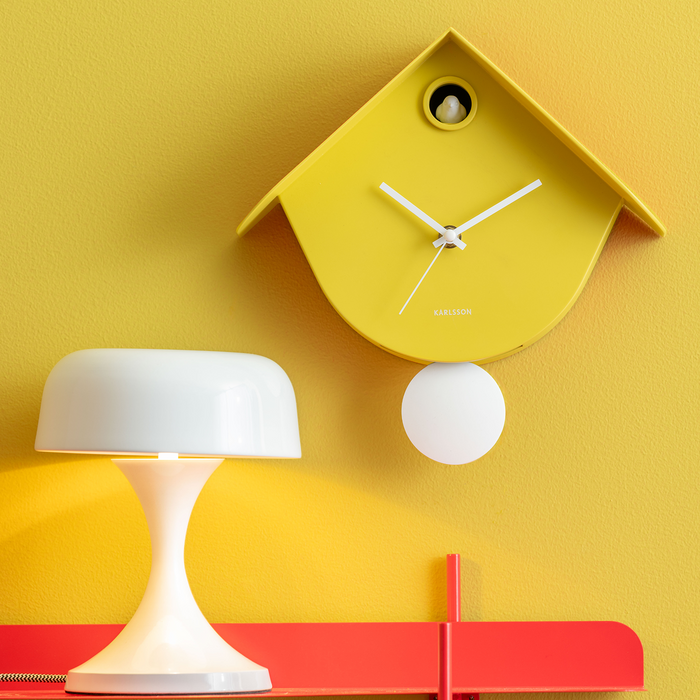 A yellow house-shaped cuckoo clock with white hands and a white hanging weight, displayed on a yellow wall above a white table lamp and a red shelf.