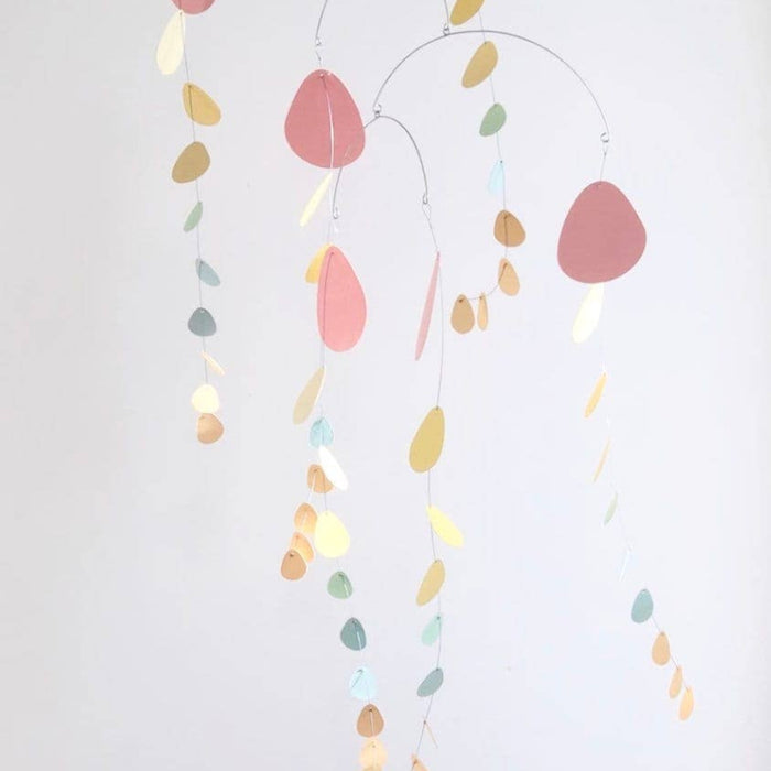 A minimalist mobile with hanging metal wires and pastel-colored teardrop-shaped ornaments in pink, gold, blue, and cream, suspended against a plain light gray background.