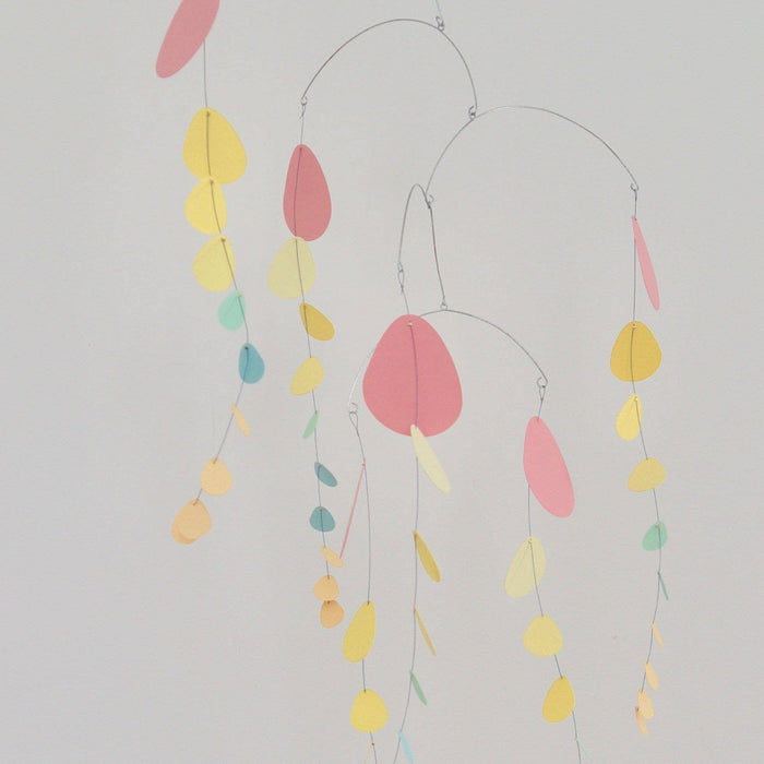 A minimalist mobile or wind spinner with thin metal hanging wires and scattered pastel paper shapes in pink, yellow, teal, and peach against a plain light background.