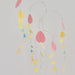 A minimalist mobile or wind spinner with thin metal hanging wires and scattered pastel paper shapes in pink, yellow, teal, and peach against a plain light background.