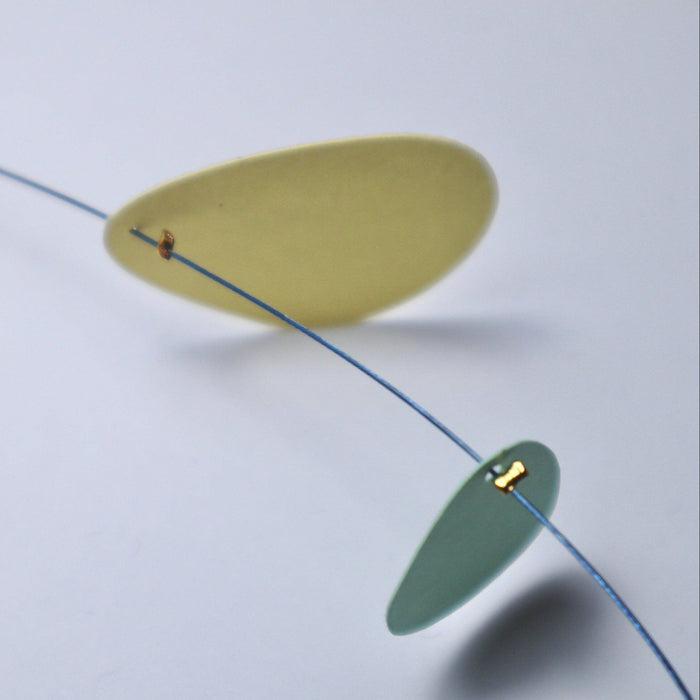A close-up of a minimalist mobile or hanging sculpture with translucent oval discs suspended on a thin blue wire against a light gray background.
