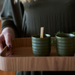 A person in a dark olive green top holds a wooden tray carrying two ribbed ceramic cups in a muted green color, with a small wooden stirrer in one cup.