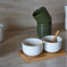 A set of ceramic tableware including white bowls with wooden spoons and dark green cups, arranged on a light wood tray against a textured beige stone background.