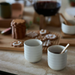Two white ribbed ceramic cups on a tabletop, one filled with a dark drink and one empty, with cookies and a wooden spoon in a softly blurred indoor setting.