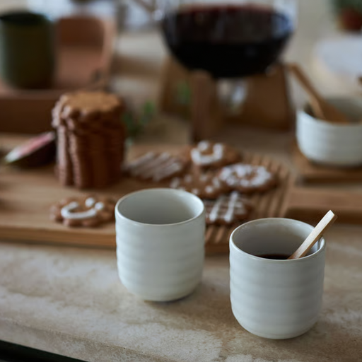 Two white ribbed ceramic cups on a tabletop, one filled with a dark drink and one empty, with cookies and a wooden spoon in a softly blurred indoor setting.