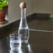Clear glass carafe and matching glass tumbler on a dark countertop, with a cork stopper resting in the neck of the carafe.