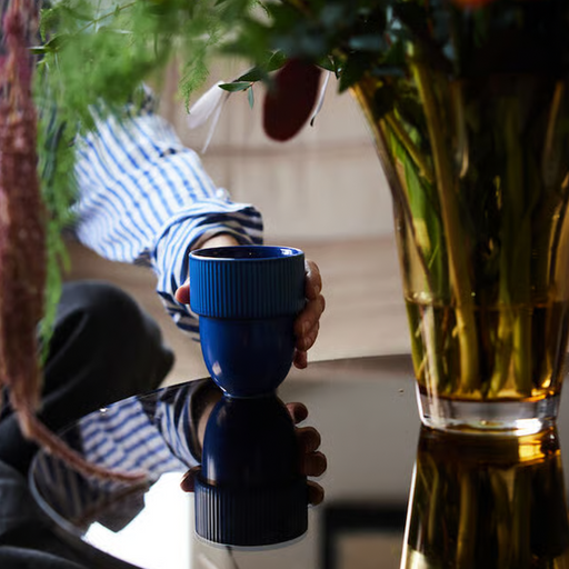 A person in a blue and white striped shirt holds a dark blue ribbed ceramic mug over a reflective black table, with a glass vase of flowers in the foreground.