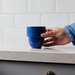 A hand wearing a denim sleeve holds a small blue cup with a ribbed top and smooth bottom, set on a speckled white countertop against a white subway tile backsplash.