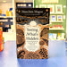 The book 'Seeing What's Hidden' by Manchán Magan stands upright on a blue table in a blurred bookstore setting. The cover features a black top section with gold Celtic-style stars and waves, a central cream-white wave pattern with a sun, and a brown bottom section with gold Celtic designs.