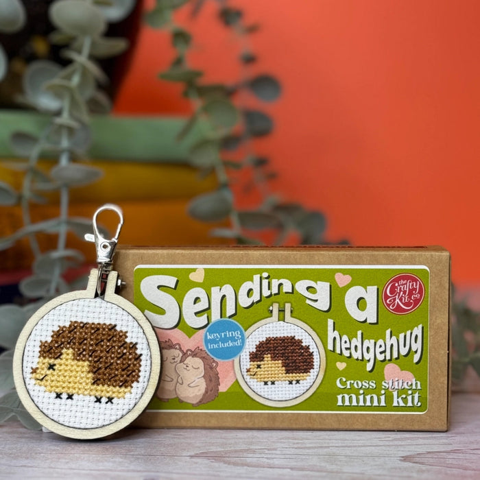 sending a hedgehug cross stitch mini kit packaging and cmbroidery hoop on a wooden surface in from of an orange background
