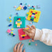 A child's hands play with colorful square pegboard toy blocks on a light blue surface, surrounded by scattered matching building pieces.