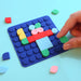A hand placing a green peg onto a colorful building block toy set on a blue square base, with loose matching pieces scattered on a light blue background.