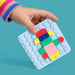 A child's hand wearing a pink, blue, and yellow striped sleeve holds a light blue square pegboard toy. The board is decorated with colorful building blocks in red, yellow, green, blue, and pink, set against a solid teal background.