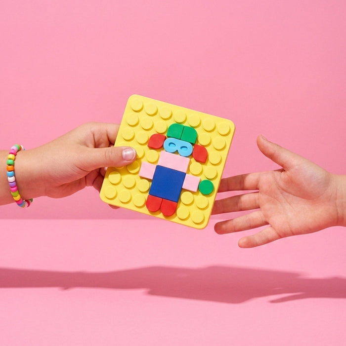 A person's hands holding a yellow square toy building-block base with a colorful assembled doll figure, set against a solid pink background.