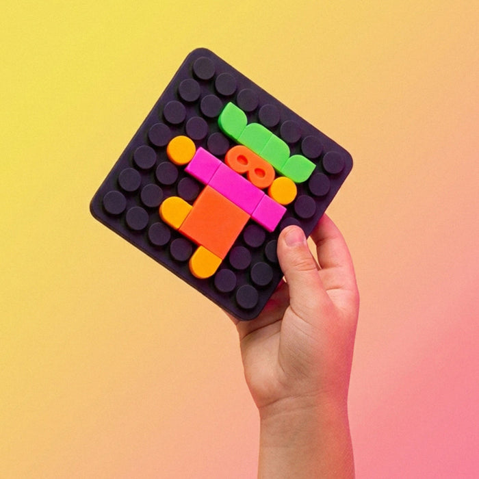 A hand holds a square black toy building block baseplate decorated with colorful plastic blocks in green, pink, orange, and yellow against a yellow-to-pink gradient background.