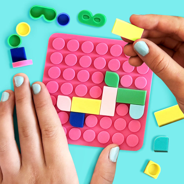 A pink silicone sensory mat with raised circular bumps, being held by two hands with pastel nail polish. Colorful plastic blocks and matching sensory pieces are scattered around on a bright turquoise background.