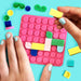 A pink silicone sensory mat with raised circular bumps, being held by two hands with pastel nail polish. Colorful plastic blocks and matching sensory pieces are scattered around on a bright turquoise background.