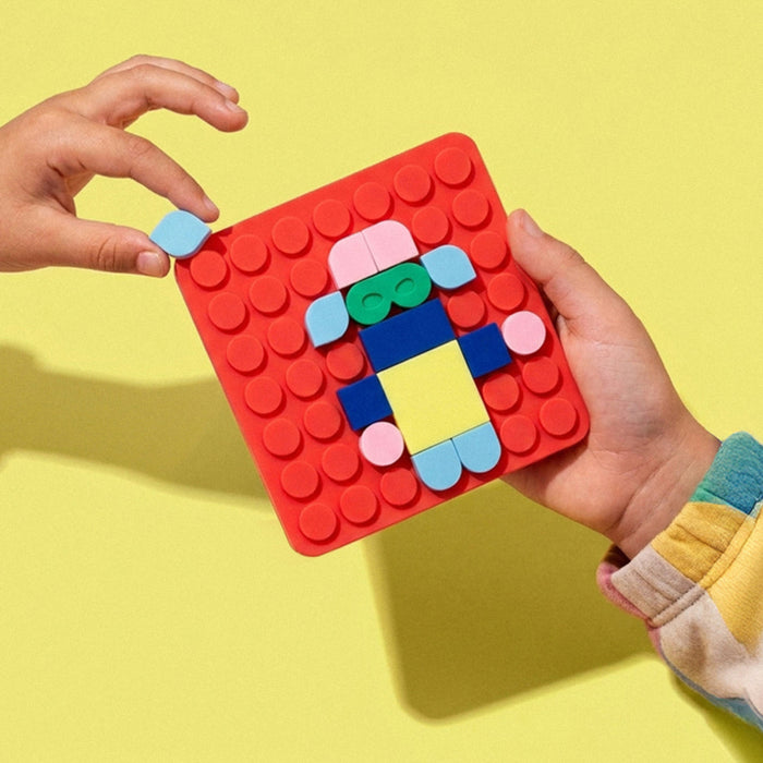 A child's hands hold a red square building block baseplate decorated with colorful toy bricks, against a solid yellow background.