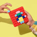 A child's hands hold a red square building block baseplate decorated with colorful toy bricks, against a solid yellow background.
