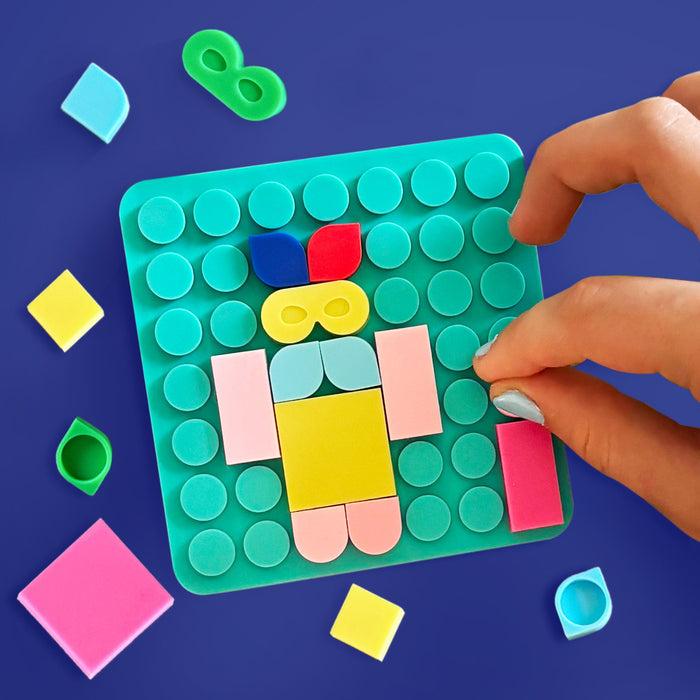 A hand is assembling a colorful geometric puzzle board on a dark blue background. The board is teal with circular studs and holds pastel pink, yellow, light blue, and red shapes, surrounded by loose matching pieces.