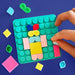 A hand is assembling a colorful geometric puzzle board on a dark blue background. The board is teal with circular studs and holds pastel pink, yellow, light blue, and red shapes, surrounded by loose matching pieces.