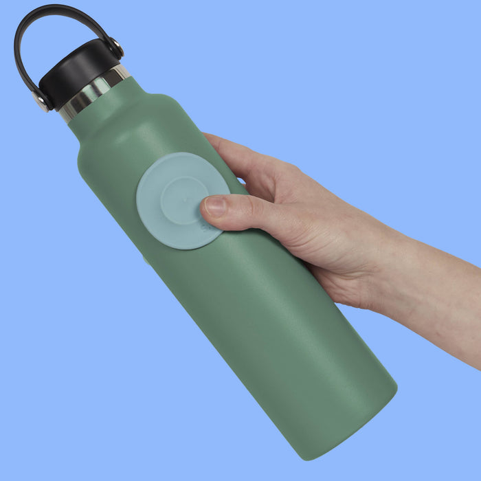 A hand holding a matte sage green insulated water bottle with a black loop handle and a light blue circular button on the front, set against a solid light blue background.