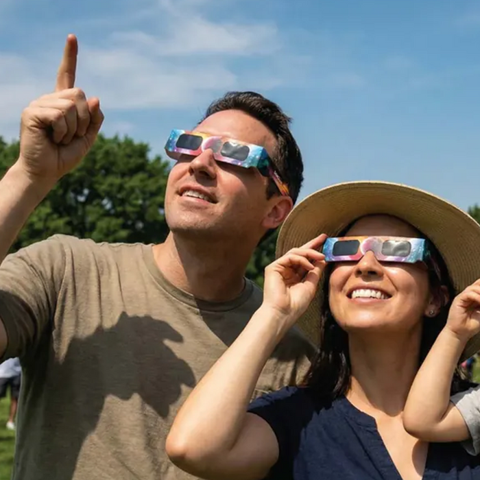 A man and a woman outdoors wearing colorful eclipse glasses under a blue sky. The man points upward, and the woman adjusts her glasses while smiling.