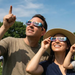 A man and a woman outdoors wearing colorful eclipse glasses under a blue sky. The man points upward, and the woman adjusts her glasses while smiling.