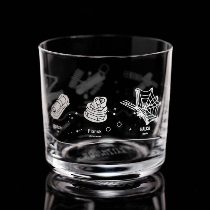 A clear glass rocks tumbler with white etched space-themed illustrations, including a rocket, a satellite dish, and a spiderweb, on a reflective black background.
