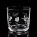 A clear glass rocks tumbler with white etched space-themed illustrations, including a rocket, a satellite dish, and a spiderweb, on a reflective black background.