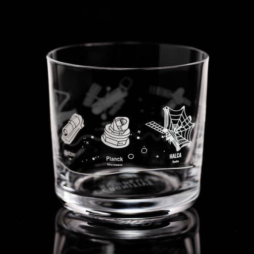 A clear glass rocks tumbler with white etched space-themed illustrations, including a rocket, a satellite dish, and a spiderweb, on a reflective black background.