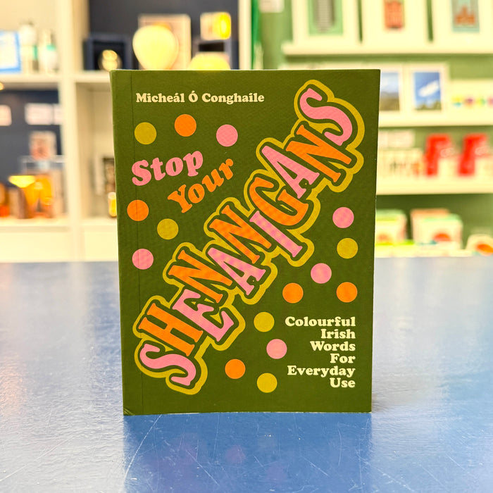 A book titled 'Stop Your Shenangans' by Micheál É Conghaile, featuring a dark olive green cover with colorful pink, orange, and yellow polka dots and bold stylized lettering, standing on a blue surface with a blurred bookstore background.