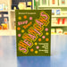 A book titled 'Stop Your Shenangans' by Micheál É Conghaile, featuring a dark olive green cover with colorful pink, orange, and yellow polka dots and bold stylized lettering, standing on a blue surface with a blurred bookstore background.