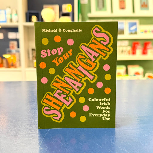 A book titled 'Stop Your Shenangans' by Micheál É Conghaile, featuring a dark olive green cover with colorful pink, orange, and yellow polka dots and bold stylized lettering, standing on a blue surface with a blurred bookstore background.