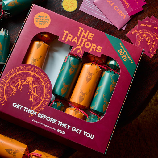 A boxed set of decorative Christmas crackers labeled 'The Traitors 2026' arranged on a dark wooden table with role cards, tickets, and additional crackers nearby.