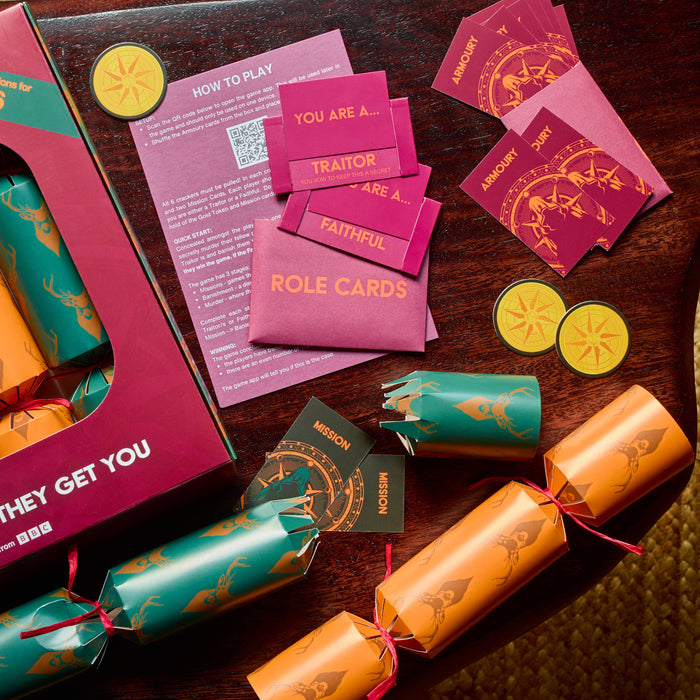 A styled flat-lay of a 'The Traitors 2026' Christmas crackers game set on a dark wooden table. The set includes a burgundy and gold box, several orange and teal wrapped crackers tied with red strings, pink role cards, a 'How to Play' guide, and yellow lemon-shaped tokens.