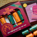 A styled flat-lay of a board game set called 'The Traitors 2026' on a dark wooden table. The set includes a deep magenta box with a clear window showing orange and teal cracker packs, several pink role cards, yellow circular tokens, and additional cracker packs tied with red strings.