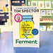 The book cover of 'Ferment' by Tim Spector is displayed upright on a blue table in a blurred bookstore setting. The cover features a white background with a large yellow and blue jar illustration and green title text.