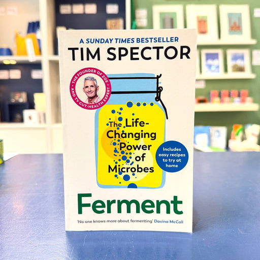 The book cover of 'Ferment' by Tim Spector is displayed upright on a blue table in a blurred bookstore setting. The cover features a white background with a large yellow and blue jar illustration and green title text.