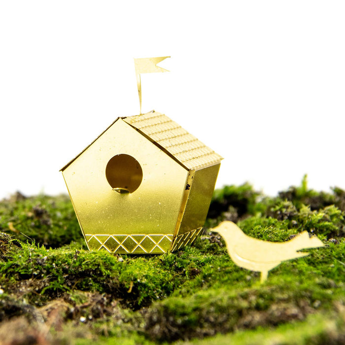 A small metallic gold birdhouse-shaped ornament with a circular entrance hole, a tiny flag on top, and a matching bird silhouette on a bed of green moss against a white background.