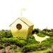 A small metallic gold birdhouse-shaped ornament with a circular entrance hole, a tiny flag on top, and a matching bird silhouette on a bed of green moss against a white background.