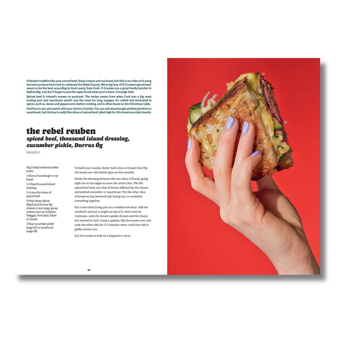 An open magazine-style spread with a recipe article about a 'rebel reuben' on the left page and a large food photo on the right page. A hand with lavender nail polish holds a thick sandwich on a bright red background.
