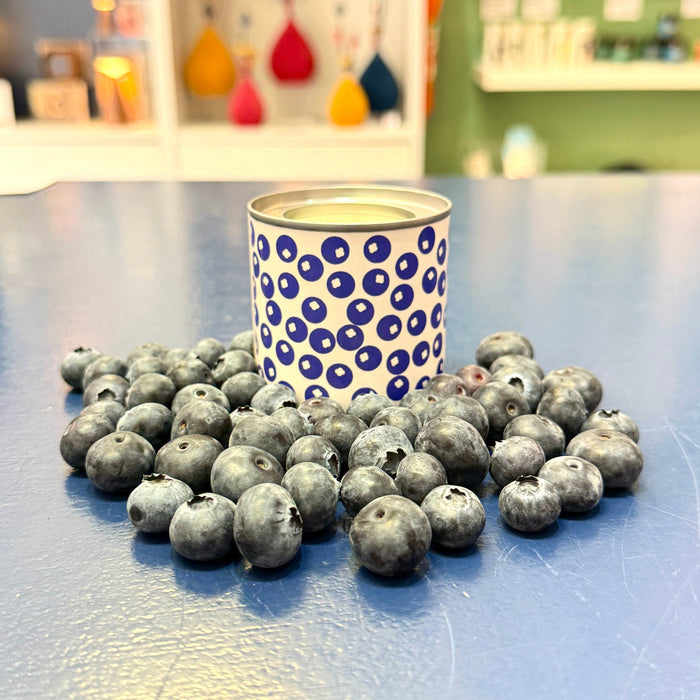 Blueberry candle tin surrounded by blueberries on a blue counter with shop background.