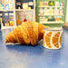 Candle tin with croissant illustrations beside a croissant on a blue counter with shop background.
