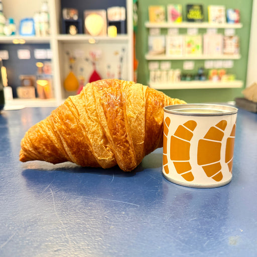 Candle tin with croissant illustrations beside a croissant on a blue counter with shop background.