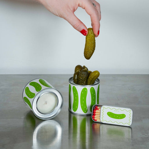 Person picking up a pickle from a can with a candle and matchbook on a reflective surface.