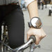 CLOSE UP OF A CIRCULAR MIRRORED BRACLET ON THE WRIST OF OF A PERSON HOLDING ONTO A BICYCLE HANDLEBAR AND WEARIING BLACK JEANS 