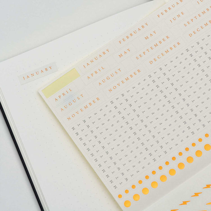 sticker page with months, letters gold dots and lightening bolts on top of a gridded page with a January sticker in top left corner