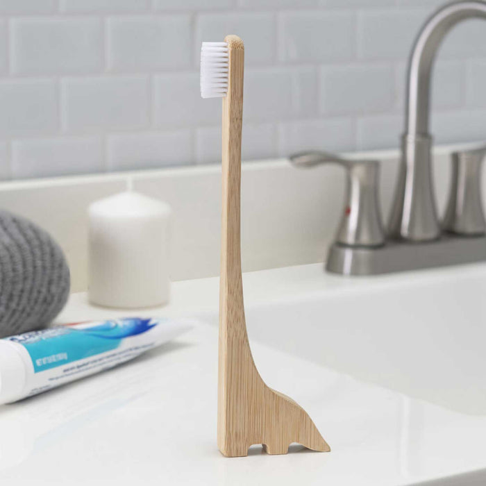 dinosaur shaped wooden toothbrush on a white sink with grey tiles and blue and white toothpaste tube